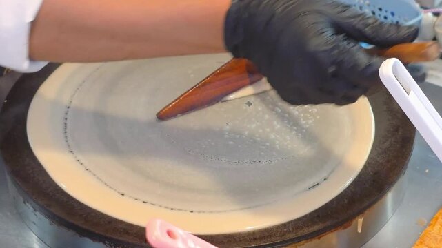 chef spreading liquid batter with wooden tool on hot circular griddle to make thin crepe at street food stall
