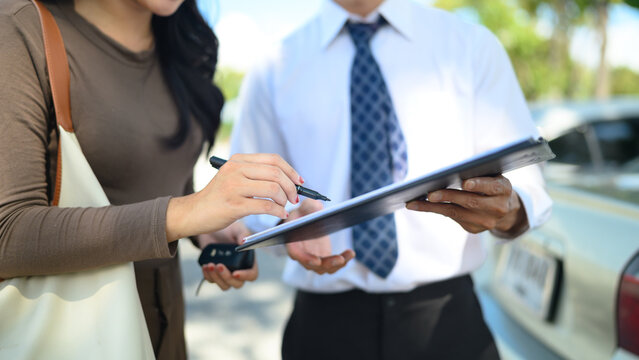 Woman signing car rental agreement on clipboard with agent holding car keys at outdoor lot