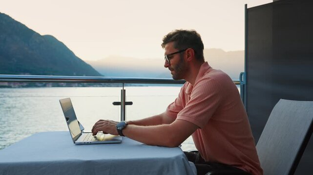 Focused remote worker using laptop on peaceful lakeside balcony at sunset, concentrating on online tasks while enjoying tranquil outdoor workspace and mountain views