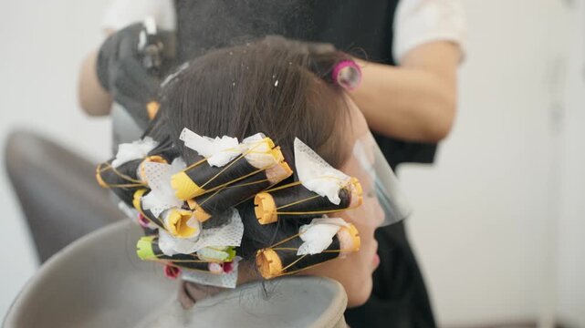 Stylist in Korean beauty salon prepares female client for perm with rolled perm rods, end papers and protective face shield while she leans back at shampoo basin