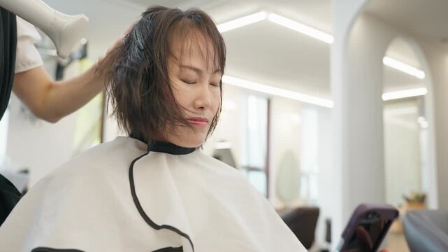 Hairstylist in modern Korean beauty salon blow dries wet short bob hair of middle aged woman wearing white cape as she looks at her smartphone