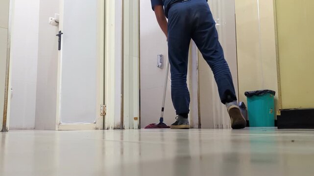 A janitor mops the tiled floor of a office restroom near an open stall, with a green waste bin and bathroom fixtures in the background.