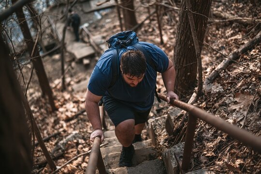 An overweight man struggles while hiking up a steep set of outdoor stairs in a wooded forest area