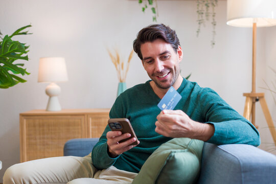 White man using smartphone and credit card sitting on sofa