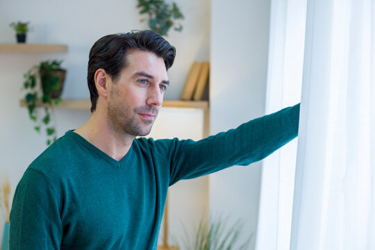 White man stands inside home looking thoughtfully out window
