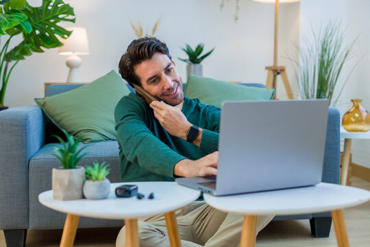 White man multitasking with laptop and phone in living room at home