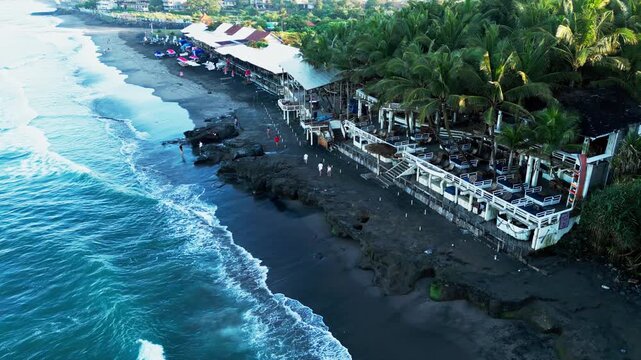 La Brisa Bali beach club aerial view at Echo Beach Canggu showing ocean waves crashing on dark volcanic sand coastline surrounded by lush tropical palm trees in sunny morning aerial drone view