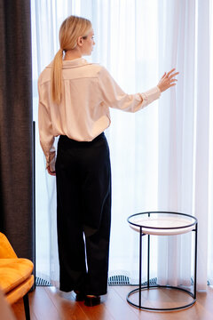 Woman standing beside bedroom curtains in a comfortable home. She reaches toward the sheer fabric at the window, with soft daylight entering the bedroom and a relaxed domestic setting around her.