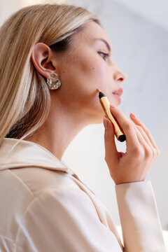 Close-up of a woman applying concealer to her cheek with a pen. Her hand holds the brush applicator beside the jawline, showing a vertical side portrait focused on precise makeup application.