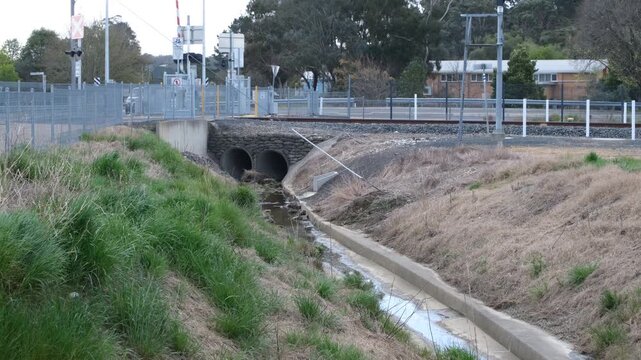 Twin stone-faced culverts and a concrete-lined drainage channel with a shallow water, passing under a railway embankment with a level crossing in the background, in Beaufort, Victoria, Australia. 