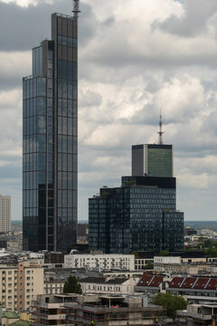Glass skyscraper rising above lower city buildings beneath a cloudy sky in the Wola district of Warsaw with layered urban architecture and distant towers