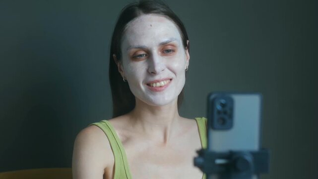 Young female influencer recording vlog on smartphone in cosmetic facial mask, sharing skincare routine, beauty tips with online followers from her home. Beauty blogger vlogging with cosmetic face mask