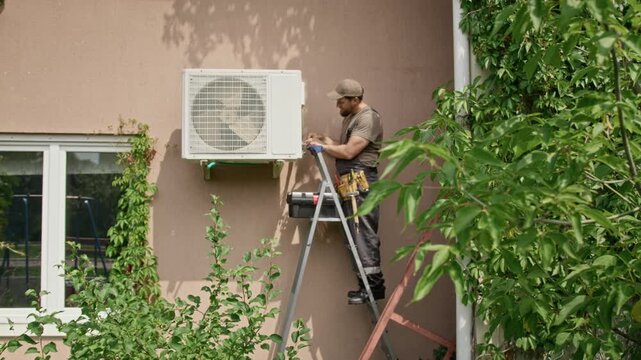 Young adult Caucasian male technician standing on ladder installing outdoor air conditioning unit on house wall. Video suitable for HVAC services and home repair campaigns