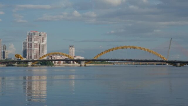 Golden arched dragon bridge spanning calm river with reflective water surface and city skyline in soft midday light - panoramic urban waterfront view, modern architecture and steady traffic movement
