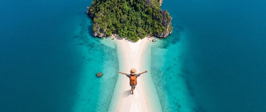 Aerial view of a female traveler with an orange backpack and straw hat standing on a narrow white sandbar beach surrounded by crystal clear turquoise ocean water toward a lush tropical island.
