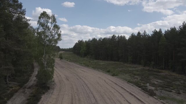 Wide aerial drone shot over a sandy dirt road running through a pine forest under a blue sky with scattered clouds near Lychen, Brandenburg, Germany.