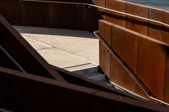 Rust colored steel walls frame a concrete staircase and walkway with angular railings in modern urban architecture viewed from above