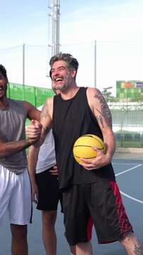 Mature man with tattoos holding a ball and laughing with his team. Mentorship and camaraderie between generations on a basketball court