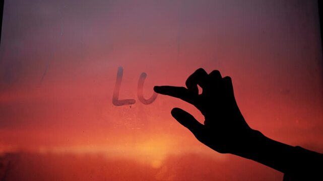Romantic silhouette of a hand tenderly writing the word love and a small heart shape on a foggy window, with the warm colors of a dramatic sunset creating a heartfelt backdrop