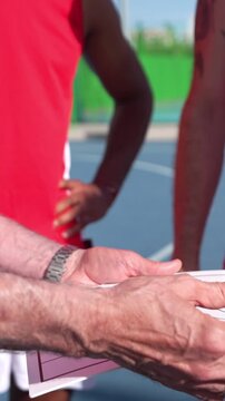 Close up of an elderly coach's hand drawing a play on a clipboard for his diverse team on a sunny outdoor court during a timeout