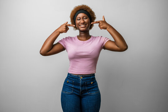 Young african american woman in pink clothes smiling and showing teeth, pointing with finger to mouth to point out dental health, cheerful studio portrait