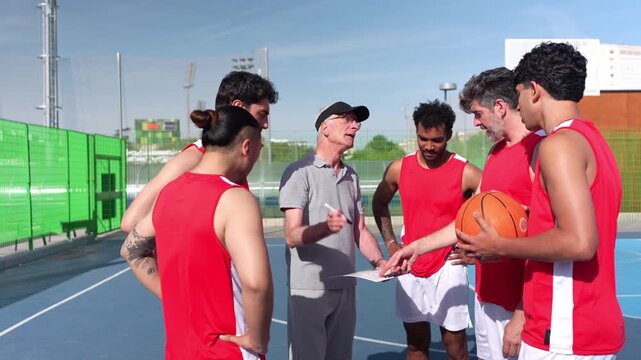 Elderly coach talking to a multiracial group of male basketball players, explaining game strategy on an outdoor court. Team celebrating together