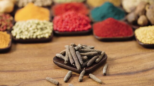 indian long pepper catkins or pippali in a square bowl on a wooden table, surrounded by various spices and herbs.