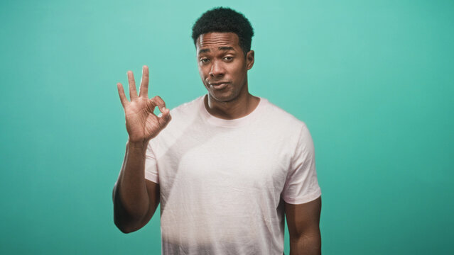 Man in pale pink t shirt making ok sign with right hand while facing camera against teal backdrop in studio; playful approval.