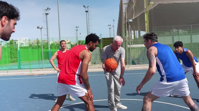 Elderly coach instructing a multicultural group of young men playing basketball. The team practices drills, scores a basket, and celebrates