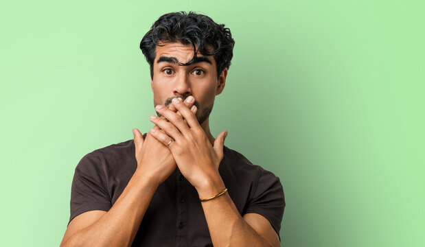 Young hispanic man casual portrait in studio covering mouth with hands to cover a mistake, covering hand in shock over a secret, shocked and surprised expression