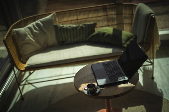 A laptop and coffee cup sit on a wooden table in a sunny office
