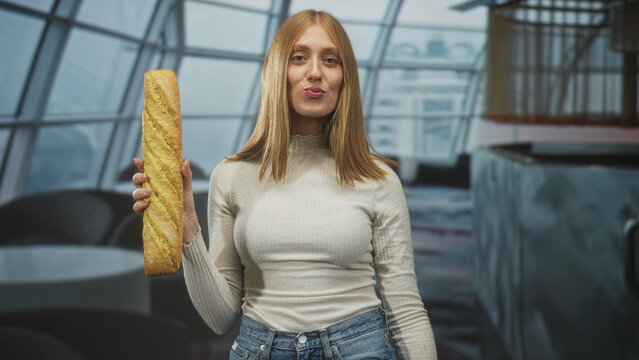 Young redhead woman holding baguette in right hand, puckered lips and casual pose in airport terminal with large windows; playful travel moment.