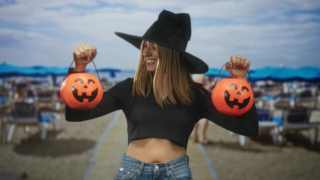 Woman holding two orange pumpkin buckets and raising arms while smiling, showing bare midriff and wearing a witch hat in studio; playful.