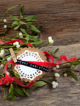 "Merry Christmas" greeting card, red ribbon and Mistletoe plant branches close up on rustic wooden background. decor for Christmas, Advent holidays, festive winter season
