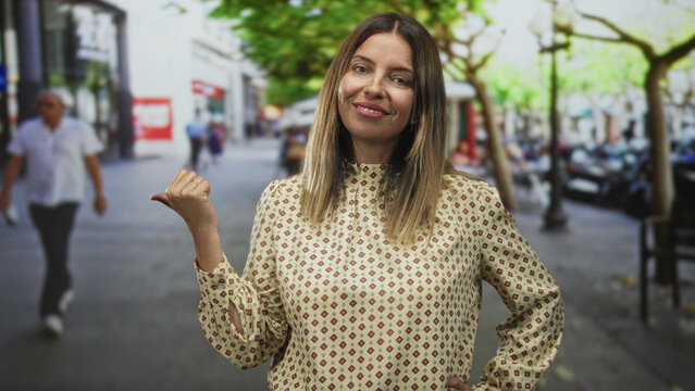 Woman pointing thumb to the side with visible hand gesture on a street, smiling with casual posture; confidence playfulness.