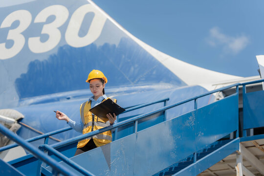 Smiling Asian female aircraft engineer wearing yellow safety helmet and vest using digital tablet under airplane wing