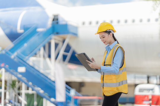 Asian female aircraft engineer wearing yellow hardhat and safety vest using digital tablet to inspect airplane jet engine.