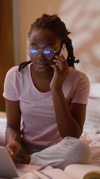 Multitasking African American business woman talking smartphone call and laptop at home bedroom night female freelancer speaking mobile phone typing computer writing notes working online communication
