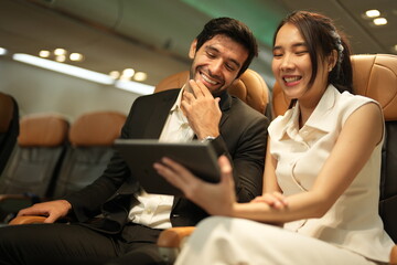 A bearded businessman and a young woman share a tablet screen in adjacent seats on board a commercial flight, good for connection and travel themes, an easy, natural moment in the air © ultramansk