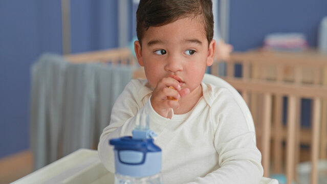 Boy holding cracker to mouth with hand in home nursery highchair beside crib, sippy cup on tray; gentle curiosity.