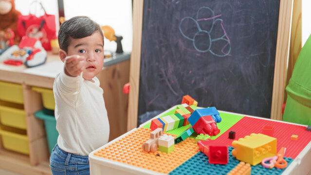 Boy pointing with outstretched hand at camera beside colorful toy blocks on a play table in a building; curiosity.
