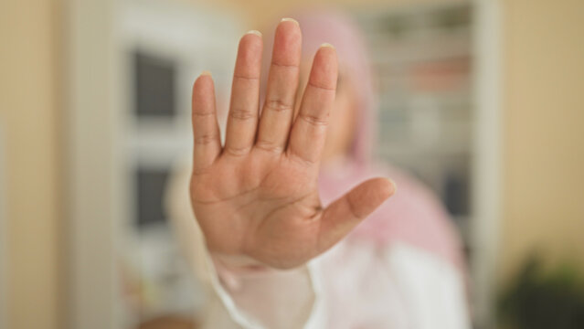 Woman palm forward stop gesture toward camera in studio, hijab wearing young arab with bookshelf and plant visible; privacy refusal.