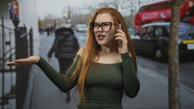 Redheaded woman on a street holding a smartphone to her ear with palm raised in an exasperated shrug, wearing glasses and green off shoulder top; frustration confrontation.