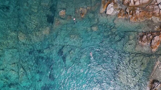 An overhead view of crystal-clear, turquoise waters along a rocky coastline, with people snorkeling.