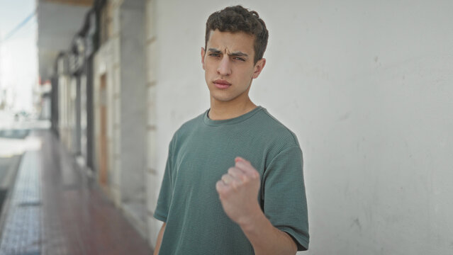 Young hispanic man with clenched fist raised and furrowed brow standing against a white wall on an urban street; defiance.