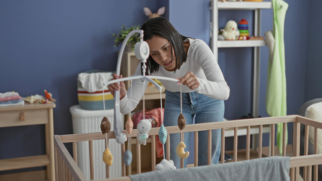 Woman adjusts a hanging baby mobile over a wooden crib, points finger to a plush toy and leans in amid nursery furniture in building; tenderness.