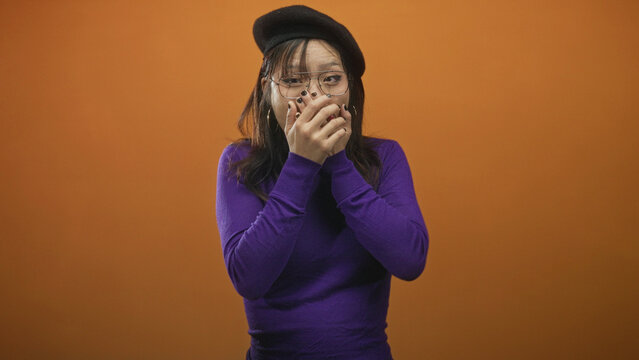 Woman covers mouth with hands and gazes wide eyed in studio wearing purple sweater, beret and glasses; surprise shyness.