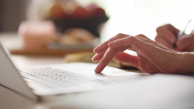 Close up of female hand using touchpad on laptop while working online. Woman browsing internet and operating computer near notebook while sitting at table in bright modern home interior.