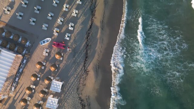 An overhead view of a sandy beach with rows of straw umbrellas, sun loungers, and rescue boats beside foaming sea waves.