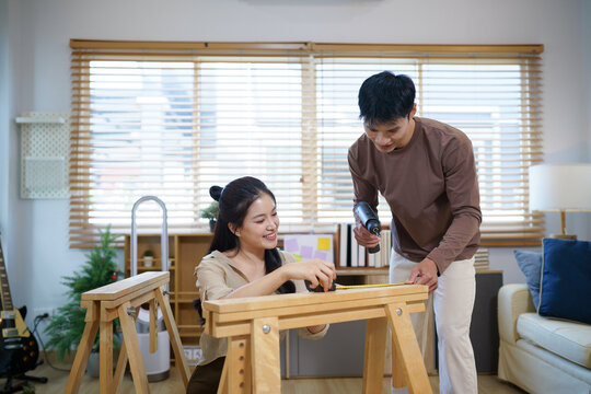 Happy Asian couple assembling furniture together at home, woman measuring wood on sawhorse while man holds power drill, ideal for lifestyle, renovation, and teamwork concepts